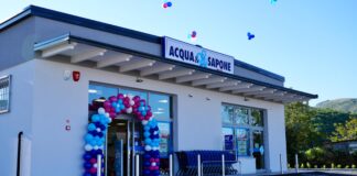 Exterior of an Acqua & Sapone store with a colorful balloon arch at the entrance and shopping carts outside, bright blue sky overhead.