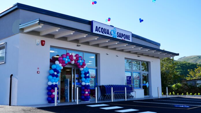 Exterior of an Acqua & Sapone store with a colorful balloon arch at the entrance and shopping carts outside, bright blue sky overhead.