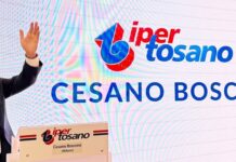 Iper Tosano rafforza la presenza in Lombardia con il nuovo store di Cesano Boscone Man in a dark suit speaks into a microphone at a podium, with a large screen displaying a logo and the words Cesano Boscone in the background