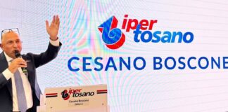 Man in a dark suit speaks into a microphone at a podium, with a large screen displaying a logo and the words Cesano Boscone in the background