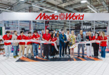 MediaWorld e Bennet consolidano la partnership con un nuovo shop-in-shop Group of people in red shirts line up behind a Media World ribbon at a store opening, indoors with bright ceiling lights and signage visible.