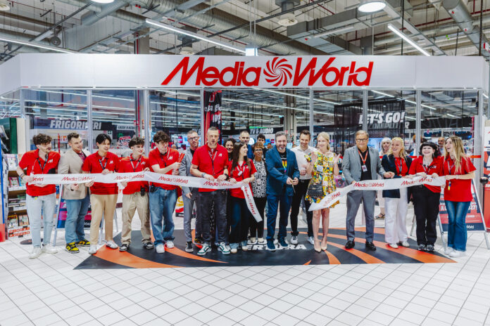 Group of people in red shirts line up behind a Media World ribbon at a store opening, indoors with bright ceiling lights and signage visible.