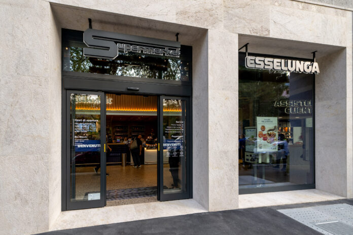 Esselunga supermarket entrance with glass doors and a 'Supermarket' sign above on a beige stone facade.
