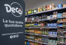 Multicedi presidia la prossimità con un nuovo Market Decò nel casertano Left promotional banner reads 'Deco La tua scelta quotidiana' with logo and social icons; shelves on the right are stocked with oils, sauces, and canned goods in a grocery store.