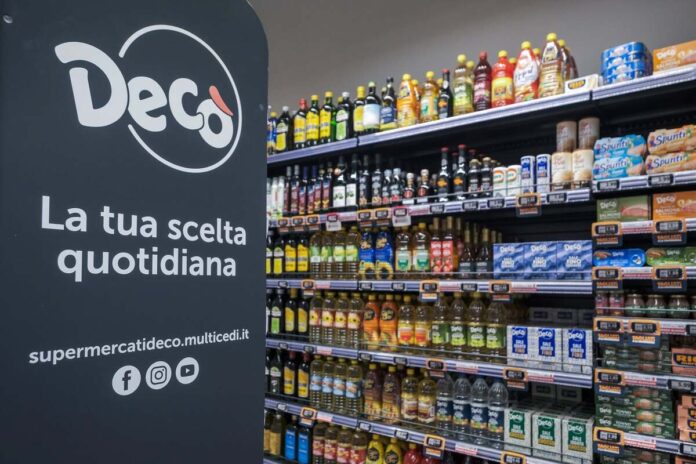 Left promotional banner reads 'Deco La tua scelta quotidiana' with logo and social icons; shelves on the right are stocked with oils, sauces, and canned goods in a grocery store.