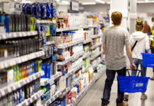 Action apre il primo punto vendita in Sicilia e porta la rete italiana a 231 negozi Back view of a man in a gray shirt pushing a blue 'ACTION' basket down a brightly lit supermarket aisle lined with shelves of products.