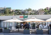 CRAI porta il format CRAIGO! nel porto di Ventimiglia Outdoor shopping area with a modern store and white patio umbrellas; a group of people in business attire chat near the entrance, hillside buildings in the background.