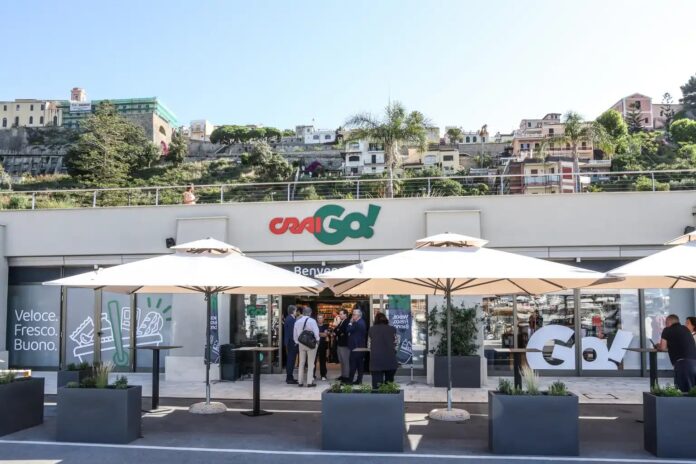 Outdoor shopping area with a modern store and white patio umbrellas; a group of people in business attire chat near the entrance, hillside buildings in the background.