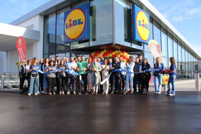 Group of people at a Lidl store during a ribbon-cutting ceremony with blue ribbon and yellow-red balloons outside the entrance.