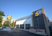 Lidl supermarket exterior with a prominent circular blue-and-yellow sign, glass storefront, and a paved parking area on a sunny day.