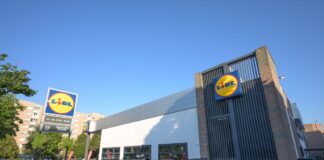 Lidl supermarket exterior with a prominent circular blue-and-yellow sign, glass storefront, and a paved parking area on a sunny day.