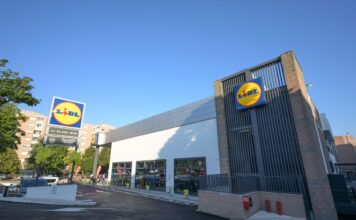Lidl supermarket exterior with a prominent circular blue-and-yellow sign, glass storefront, and a paved parking area on a sunny day.