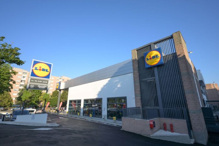Lidl supermarket exterior with a prominent circular blue-and-yellow sign, glass storefront, and a paved parking area on a sunny day.
