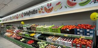 Produce aisle in a supermarket with crates of tomatoes, peppers, and other fresh fruit on display under an 'Ortofrutta' sign.