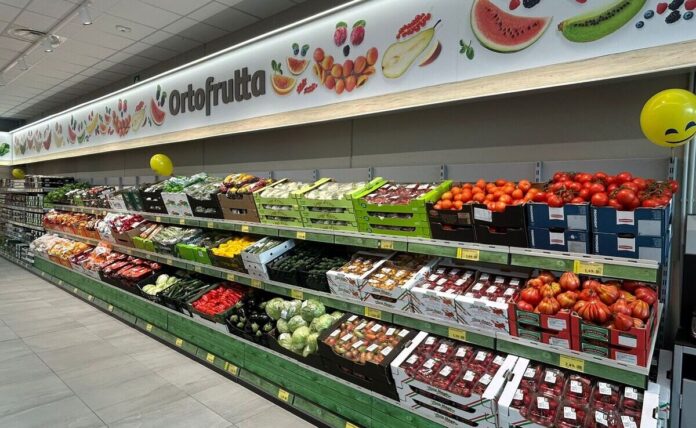 Produce aisle in a supermarket with crates of tomatoes, peppers, and other fresh fruit on display under an 'Ortofrutta' sign.