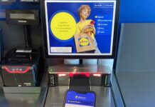 Self-checkout kiosk screen showing Lidl promotion and mobile payment options, with a hand holding a red smartphone and a Lidl bag in the foreground.