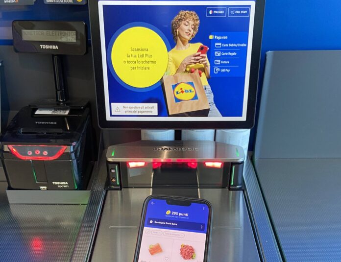 Self-checkout kiosk screen showing Lidl promotion and mobile payment options, with a hand holding a red smartphone and a Lidl bag in the foreground.