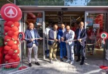 Group of men in suits cutting a long ribbon at a store opening, with red balloon column on the left and Italian flag colors in the ribbon inside the doorway.