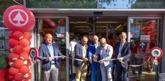 Group of men in suits cutting a long ribbon at a store opening, with red balloon column on the left and Italian flag colors in the ribbon inside the doorway.