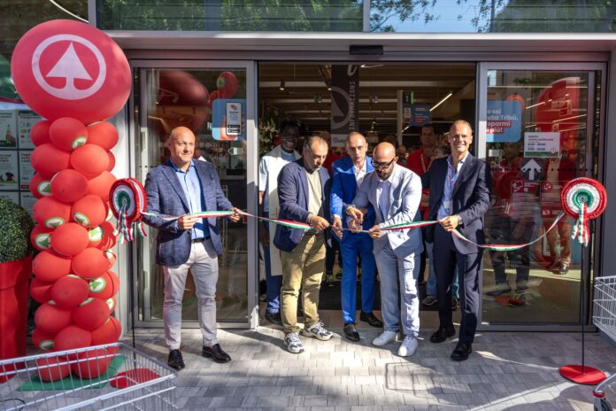 Group of men in suits cutting a long ribbon at a store opening, with red balloon column on the left and Italian flag colors in the ribbon inside the doorway.