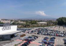 Gruppo Arena rigenera l’ex Auchan di Catania: nasce il nuovo polo commerciale Wide aerial view of a large parking lot in front of L'Arena di Catania, with a distant cityscape and hills on the horizon.