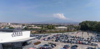 Wide aerial view of a large parking lot in front of L'Arena di Catania, with a distant cityscape and hills on the horizon.