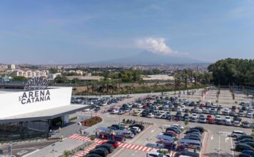 Wide aerial view of a large parking lot in front of L'Arena di Catania, with a distant cityscape and hills on the horizon.