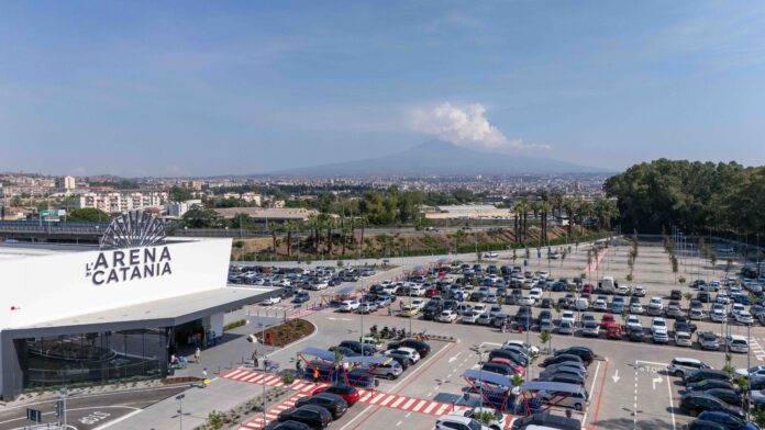 Wide aerial view of a large parking lot in front of L'Arena di Catania, with a distant cityscape and hills on the horizon.