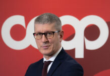 Professional man in a dark navy suit with a burgundy tie and glasses, arms crossed, against a red backdrop.