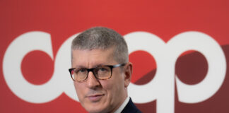 Professional man in a dark navy suit with a burgundy tie and glasses, arms crossed, against a red backdrop.