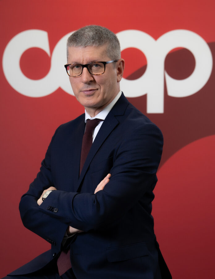 Professional man in a dark navy suit with a burgundy tie and glasses, arms crossed, against a red backdrop.