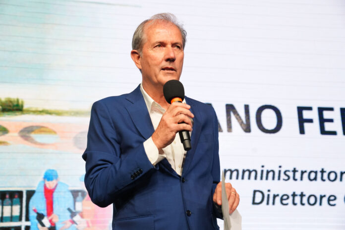 Man in a blue suit speaks into a handheld microphone on stage, background shows text and graphics behind him.