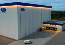 Large white industrial warehouse with blue trim and loading docks; several trailers at the dock and a red truck nearby.