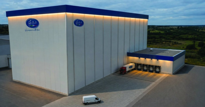 Large white industrial warehouse with blue trim and loading docks; several trailers at the dock and a red truck nearby.