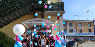 Group of people at a ribbon-cutting ceremony under blue, pink, and white balloons on a pink carpet outside a building.