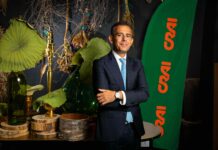CRAI SECOM cresce del 38% e consolida il ruolo di piattaforma industriale Professional portrait: man in a navy suit and blue tie stands with arms crossed beside a green banner with orange text, among botanical décor and brass items.