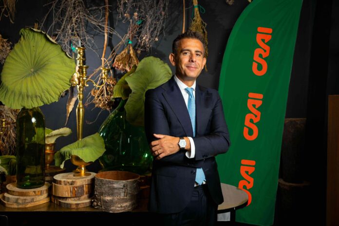 Professional portrait: man in a navy suit and blue tie stands with arms crossed beside a green banner with orange text, among botanical décor and brass items.