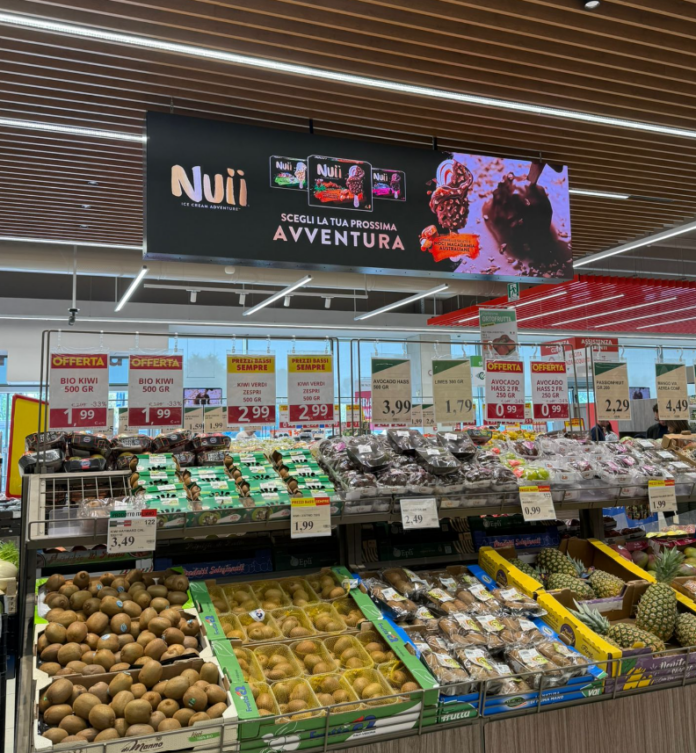 Produce aisle in a supermarket showing kiwis, kiwifruit, avocados and pineapples with a Nuii ice cream ad banner overhead.