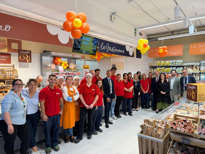 IMG-20260611-WA0001 Group of supermarket staff and managers posing for a photo under orange and yellow balloons near deli counters and signage.