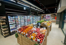 Fresh fruit display in wooden crates at a modern supermarket, with glass-door refrigerated sections behind it.