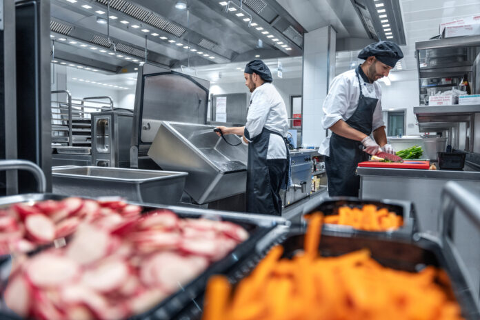 Two chefs in white uniforms and black aprons prep vegetables in a commercial kitchen; foreground shows sliced radishes and diced carrots on the prep station.