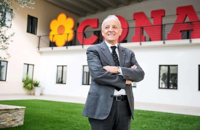 Older man in a gray suit stands with arms crossed outside a modern building with large red signage and a yellow flower logo to the left.
