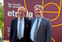 Two men in suits pose together outdoors in front of a maroon event banner, smiling for the photo.