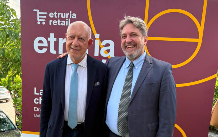 Two men in suits pose together outdoors in front of a maroon event banner, smiling for the photo.