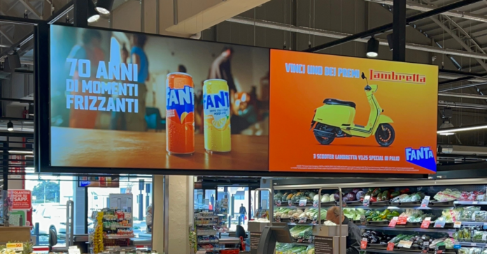 Store interior with a large double-panel advertisement: left shows Fanta cans, right shows a yellow scooter on an orange background.