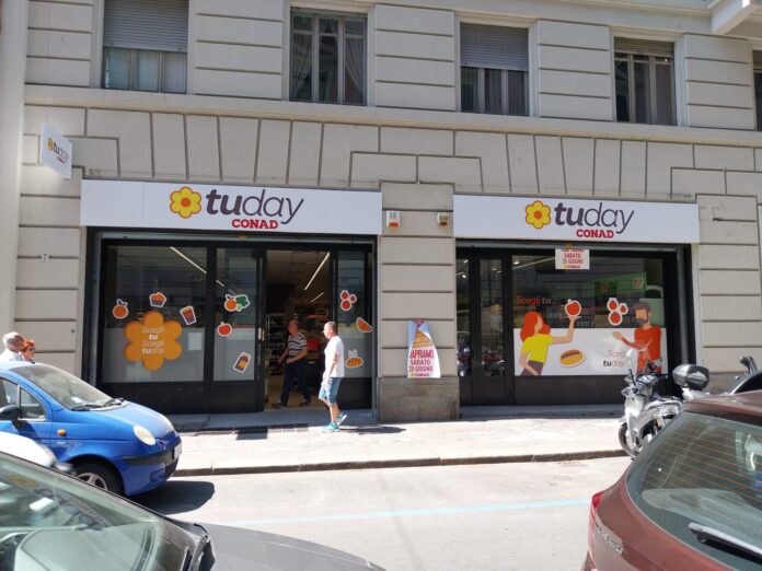 Storefront of a Today CONAD supermarket with large signs and window decals showing fruit and food icons on beige building façade