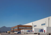Industrial warehouse with loading docks and ACT-labeled trailers parked outside under a clear blue sky, mountains in the distance.
