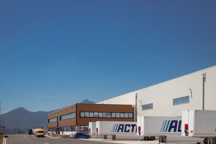 unnamed (1) Industrial warehouse with loading docks and ACT-labeled trailers parked outside under a clear blue sky, mountains in the distance.