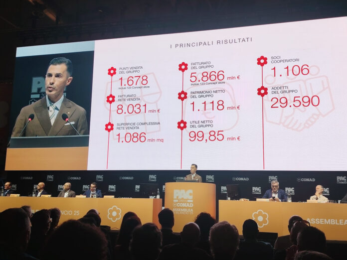 Speaker at a podium during a conference, with a large slide showing 