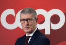Coop Italia apre una nuova fase tra bilancio solido e cambio al vertice Portrait of a man in a dark suit and glasses, standing in front of a red backdrop with large white letters behind him.